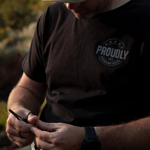 A person wearing the Proudly USA Tattered Flag Mens Cotton T-Shirt (Black) sits outdoors, focused on an object in their hands. Their face is partially hidden by a hat.