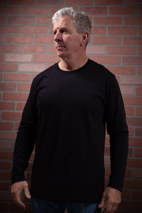An older man with short gray hair, wearing the Proudly USA Mens Long Sleeve Black Cotton T-Shirt and jeans, stands before a red brick wall, looking slightly to the side with a neutral expression.