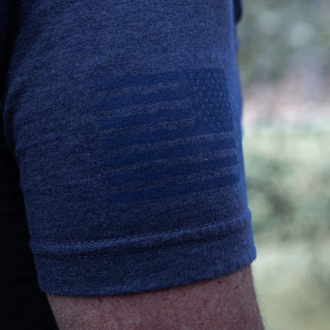 Close-up of a persons upper arm wearing the Proudly USA - Mens TriBlend T-Shirt in Heather Denim, featuring subtle patterns. The brand Proudly USA stands out against a blurred greenery background.