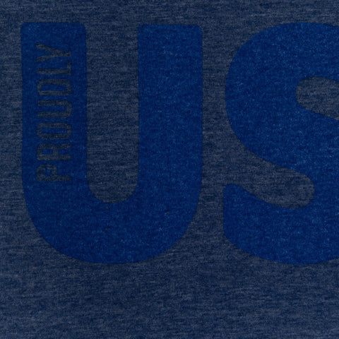 A close-up of the Proudly USA Mens TriBlend T-Shirt in Heather Denim shows large U and S letters with PROUDLY printed vertically by the U, highlighting this patriotic design from Proudly USA.