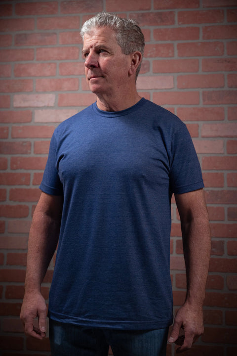 An older man with gray hair stands before a brick wall in jeans and the Proudly USA Mens Heather Denim Crewneck TriBlend T-Shirt, gazing slightly to the side with a neutral expression and his hands resting by his sides.