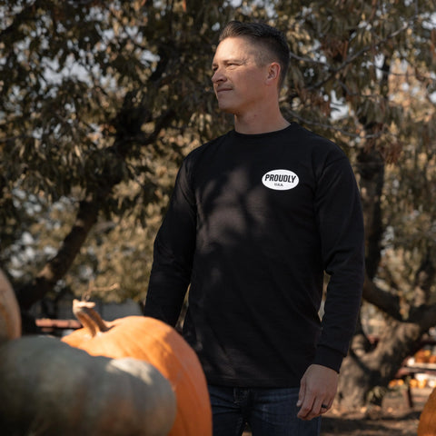 A person wearing a black Proudly USA Oval Logo Mens Long Sleeve Heavyweight Cotton T-Shirt stands outdoors among pumpkins, with trees and sunlight in the background.