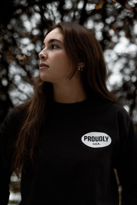 A woman with long brown hair looks to the side, wearing the Proudly USA Oval Logo Mens Long Sleeve Heavyweight Cotton T-Shirt in black. A white oval patch with the brand name is visible, with blurred branches and leaves in the background.
