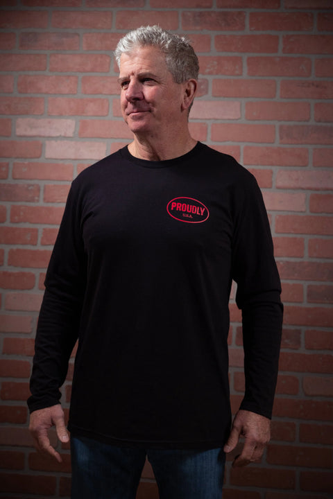 An older man with gray hair stands by a red brick wall, smiling to the side while wearing blue jeans and the Proudly USA Oval Logo Mens Long Sleeve Cotton T-Shirt (Black) with a red logo on the left chest.