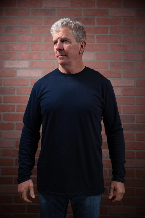 A man with short gray hair wears a Proudly USA Mens Long Sleeve Navy Cotton T-Shirt and jeans, standing against a brick wall and looking slightly left with a neutral expression.
