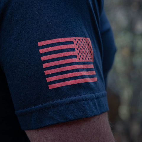 A close-up shows a persons arm in the Proudly USA Oval Logo Mens Cotton T-Shirt (Navy), made in the USA with a red, stylized American flag graphic on the sleeve. The blurred outdoor background highlights this patriotic tee.