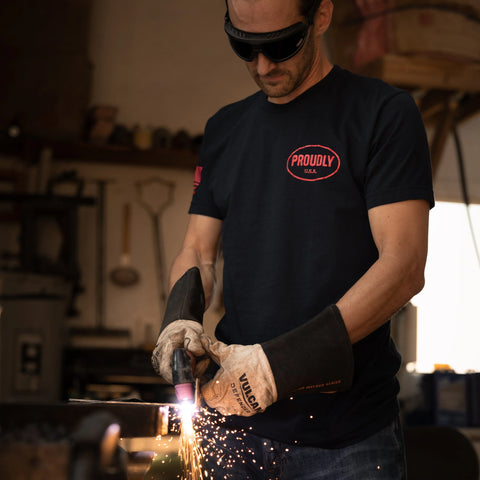 Wearing safety glasses and gloves, a person welds in a workshop with tools on the wall. They wear the Proudly USA Oval Logo Mens Cotton T-Shirt in navy, featuring a red logo. Bright sparks fly as they work.