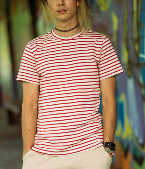 A person with long hair wears the Spiritex Sustainable Apparel Mens Striped Short Sleeve Crewneck Tee, a black choker, bracelets, and a smartwatch while standing with hands in pockets in front of a colorful graffiti wall.