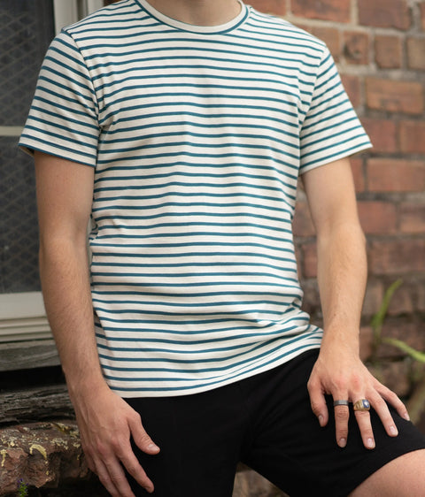 A person wearing a Spiritex | Sustainable Apparel Mens Striped Short Sleeve Crewneck Tee with green stripes and black shorts sits on a ledge by a brick wall, resting one hand on their knee.