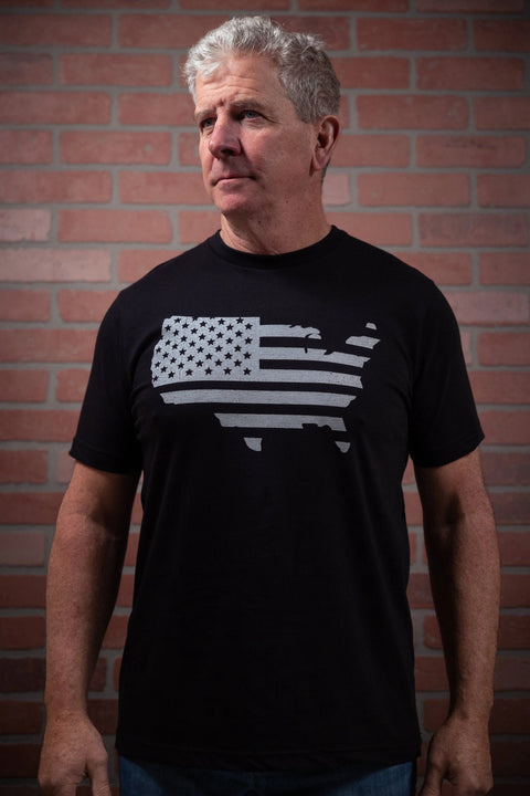 A middle-aged man with short gray hair stands by a brick wall, wearing the Proudly USA Land That I Love mens black cotton T-shirt featuring a U.S. map with the American flag design.