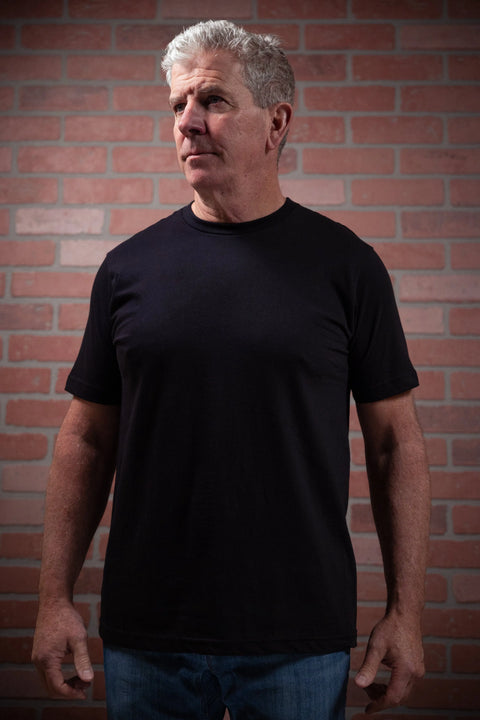 An older man with short gray hair stands before a brick wall, wearing the Proudly USA Mens Black Crewneck Cotton T-Shirt and blue jeans, gazing to the side with a serious look.