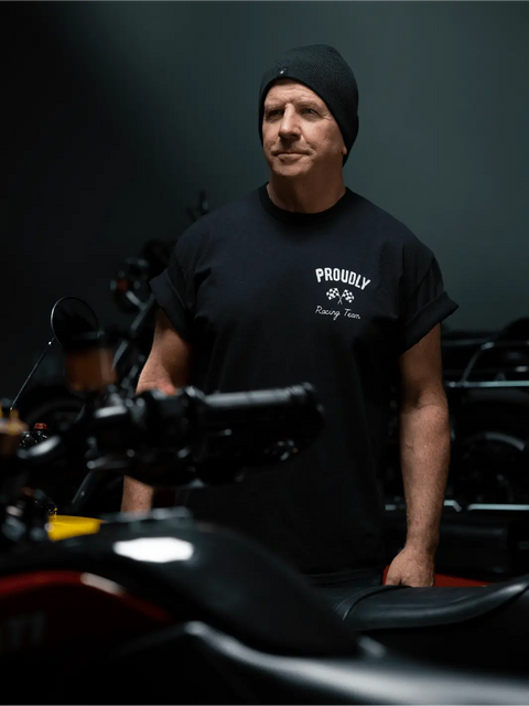 A man wearing a Proudly USA Racing Team Mens Cotton T-Shirt (Black) and a black beanie stands in a dimly lit room with motorcycles in the background.