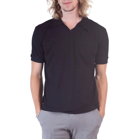 A person with long blond hair models the Spiritex | Sustainable Apparel Short Sleeve Mt. Mitchell Collared Shirt in black, paired with light gray pants, standing hands-in-pockets against a white background.