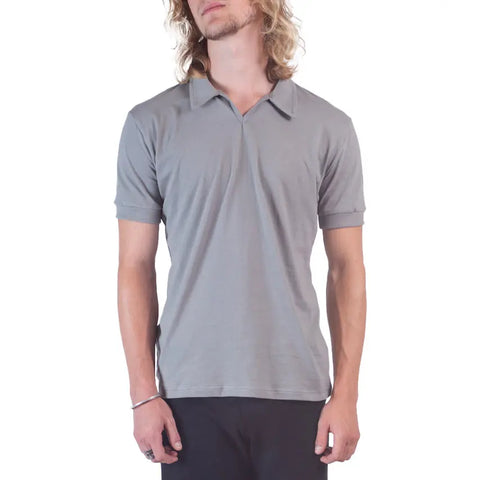 A person with long, wavy blond hair wears the Spiritex Short Sleeve Mt. Mitchell Collared Shirt in light gray organic cotton and dark pants, standing against a white background and looking slightly to the side.