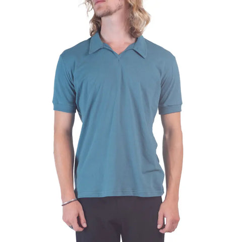 A person with long, wavy blond hair stands against a white background, wearing the Spiritex | Sustainable Apparel Short Sleeve Mt. Mitchell Collared Shirt in teal and black pants, face turned slightly to the side.