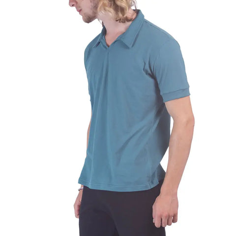 A person with light skin and long blonde hair is wearing the Spiritex Short Sleeve Mt. Mitchell Collared Shirt in teal, made in the USA, paired with black pants, standing against a plain white background facing slightly to the side.