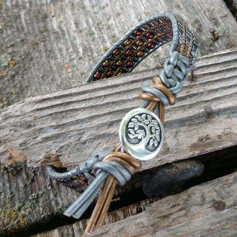 The Mens Tree of Life Intricately Beaded Leather Wrap Bracelet by Alexa Martha Designs features brown and gray leather, earth-tone beads, and a Tree of Life button, beautifully displayed on rustic wooden planks.