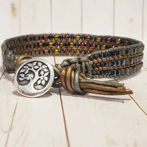 The Mens Tree of Life Intricately Beaded Leather Wrap Bracelet by Alexa Martha Designs features earth-tone brown and gold beads, a silver Tree of Life button closure, and is displayed on a light wood surface.
