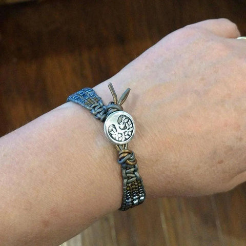 A close-up of a hand wearing Alexa Martha Designs Mens Tree of Life Intricately Beaded Leather Wrap Bracelet, featuring blue and tan threads and a round Tree of Life button closure. Wooden flooring in the background enhances its earthy charm.
