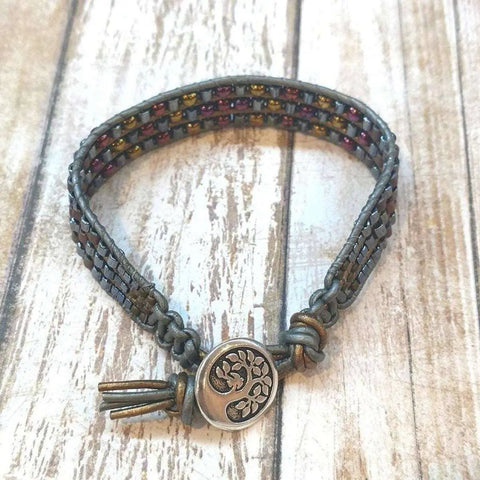The Mens Tree of Life Intricately Beaded Leather Wrap Bracelet by Alexa Martha Designs features multicolored beads on grey leather with a round tree-engraved button clasp, shown on a wooden surface.