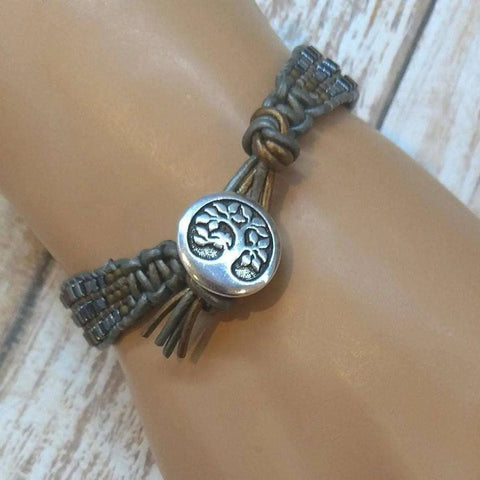 The Mens Tree of Life Intricately Beaded Leather Wrap Bracelet by Alexa Martha Designs features a silver tree of life charm and is shown worn on a wrist against a light wooden background.
