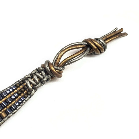 A close-up of the Mens Tree of Life Intricately Beaded Leather Wrap Bracelet by Alexa Martha Designs, featuring a metallic cord, detailed silver and bronze knot, blue beads, and looped clasp against a plain white background.