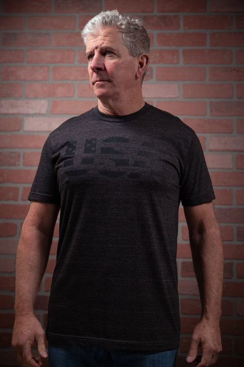 An older man with short gray hair, wearing jeans and a Proudly USA Patriotic USA Mens TriBlend T-Shirt (Heather Charcoal), stands in front of a brick wall, gazing slightly to the side with a neutral expression.