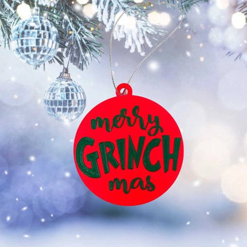 The Chatelet Manufacturing Merry Grinchmas Christmas Ornament—Decorative Holiday Ornament, Made in the USA—hangs on a snowy branch beside a silver bauble, adding festive cheer with its vibrant design against a wintry backdrop.