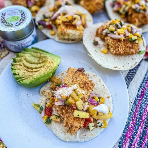 A plate with a sliced avocado and a taco filled with crispy breaded fish, corn salsa, and sour cream. More tacos and a Gustus Vitae Mexican Seasoning Gift Set - Tastes of Mexico artisanal spice blends six pack are in the background.