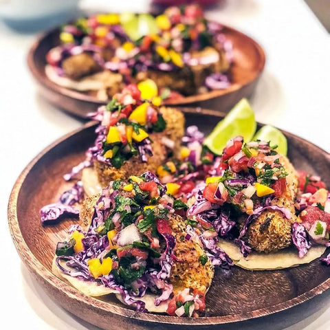 Two wooden plates hold tacos topped with purple cabbage slaw, tomatoes, yellow bell peppers, onions, and fresh herbs. Each taco is seasoned with Gustus Vitae’s Mexican Seasoning Gift Set and served with lime wedges for garnish.