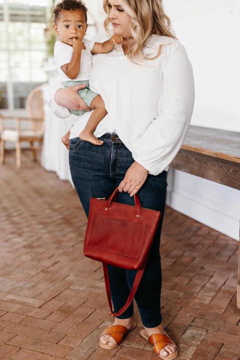 A woman in jeans and a white blouse holds a toddler on her hip and carries the Urban Southern Quinn Crossbody. She wears brown sandals, and they stand indoors on a brick floor near a wooden table.