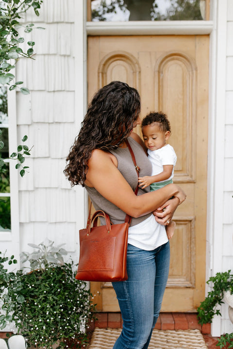 A woman with curly hair holds a young child outside a house with potted plants, wearing jeans, a sleeveless top, and carrying the Urban Southern Quinn Crossbody bag.