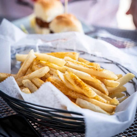 A basket of French fries, seasoned with Gustus Vitaes Midwestern Classic Seasonings Collection, rests on white parchment atop a metal mesh table, while two small sandwiches appear blurred in the background.