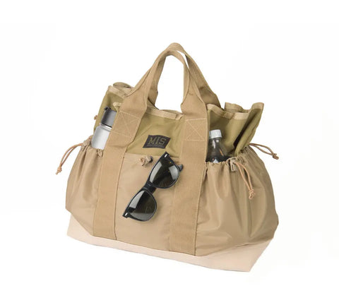 The MIS Multi Pocket Tote Bag in Coyote Tan, made in the USA from water-resistant Cordura nylon, features multiple pockets, two handles, a front logo patch, and sunglasses clipped to the front. It holds a silver water bottle and a clear bottle.