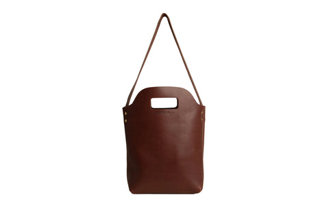The Urban Southern Mini Market Tote, a brown leather handbag with a rectangular cut-out handle and long strap, is American-made and shown against a white background.