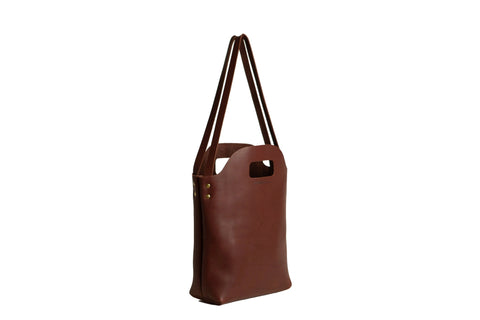 The Urban Southern Mini Market Tote is a brown leather handbag with a cutout handle, two long shoulder straps, and brass rivet accents—American-made and shown upright on a white background.