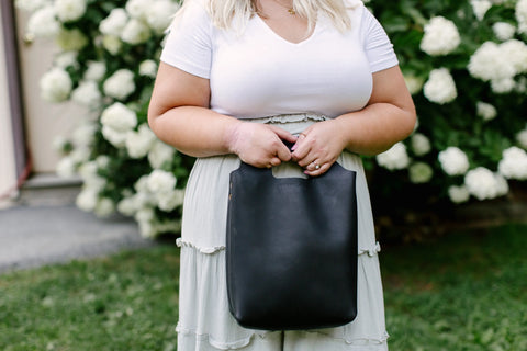 A woman in a white top and light skirt stands outdoors holding the Urban Southern Mini Market Tote, with blooming white flowers and greenery in the background.