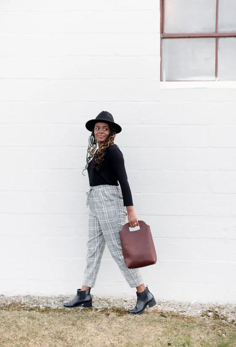 A woman in a black hat, black top, plaid pants, and black boots walks outside along a white brick wall, carrying the Urban Southern Mini Market Tote by her side.