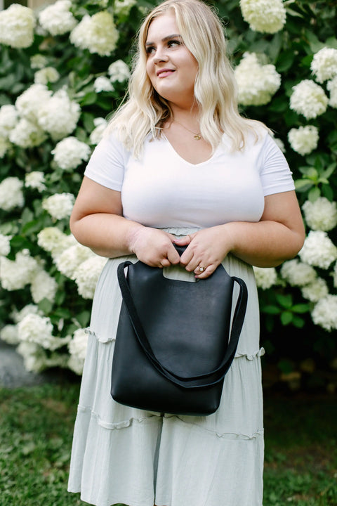 A woman with blonde hair in a white top and light skirt stands outdoors by white hydrangeas, holding an Urban Southern Mini Market Tote and smiling slightly to the side.