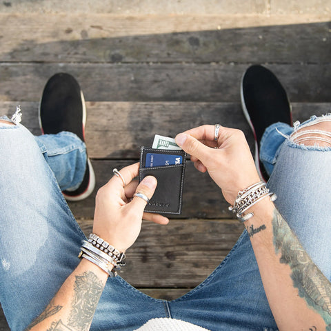 A tattooed person with bracelets, ripped jeans, and black slip-ons sits on wood, holding a CASUPO Minimalist Wallet - Black with cards and cash. The top-down photo highlights the sleek wallet design.