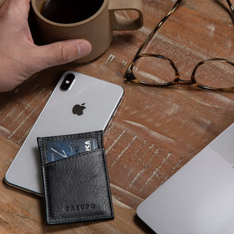 A hand holds a coffee cup beside an iPhone, eyeglasses, the CASUPO Minimalist Wallet - Black with credit cards, and a silver laptop on a wooden table.