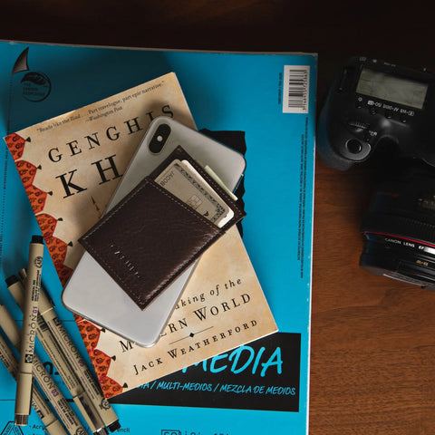 A CASUPO Minimalist Wallet - Black with cash sits atop "Genghis Khan" and a blue book, surrounded by pens and a DSLR camera, all arranged on a wooden surface.