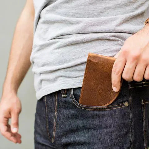 A person in a gray shirt and dark jeans is putting a Rogue Industries Rogue Front Pocket Wallet - Classic Made in Maine Edition into their front jeans pocket. Only their torso, arm, and hand are visible.