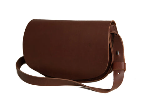 The Urban Southern Half Moon Crossbody is a minimalist brown shoulder bag made from full-grain leather, featuring a flap closure and adjustable strap—American-made and shown on a white background.
