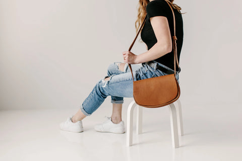 A person in a black top, ripped blue jeans, and white sneakers sits sideways on a white chair, holding the Urban Southern Half Moon Crossbody full-grain leather bag against a plain white background.