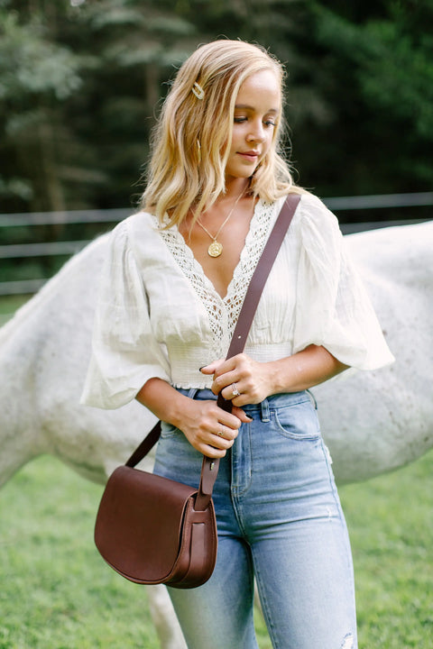 A blonde woman in a white blouse and blue jeans stands outdoors by a white horse, holding the Urban Southern Half Moon Crossbody. She looks down with greenery and a fence in the background.