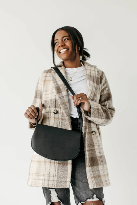 A woman with dark hair pulled back smiles while holding the Urban Southern Half Moon Crossbody, an American-made leather bag. She wears a plaid coat and white top, standing against a plain, light background.