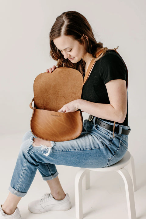 A woman in a black shirt and ripped jeans sits on a white stool, looking into an open Urban Southern Half Moon Crossbody bag on her lap against a plain white background.