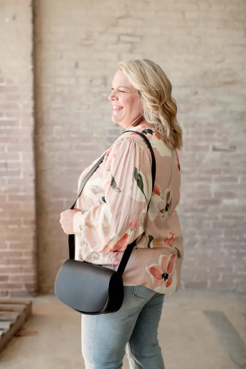 A smiling blonde woman, wearing a floral blouse and light blue jeans, carries the Urban Southern Half Moon Crossbody bag. She stands indoors by a light brick wall and pallet, facing sideways.