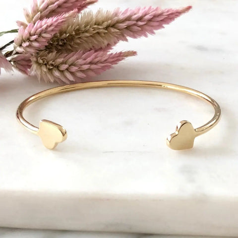 The GOLD VALEN BRACELET by Sarah Cecelia, featuring heart-shaped ends, rests on white marble with pink dried flowers—an elegant minimalist jewelry piece and a perfect gift for her.